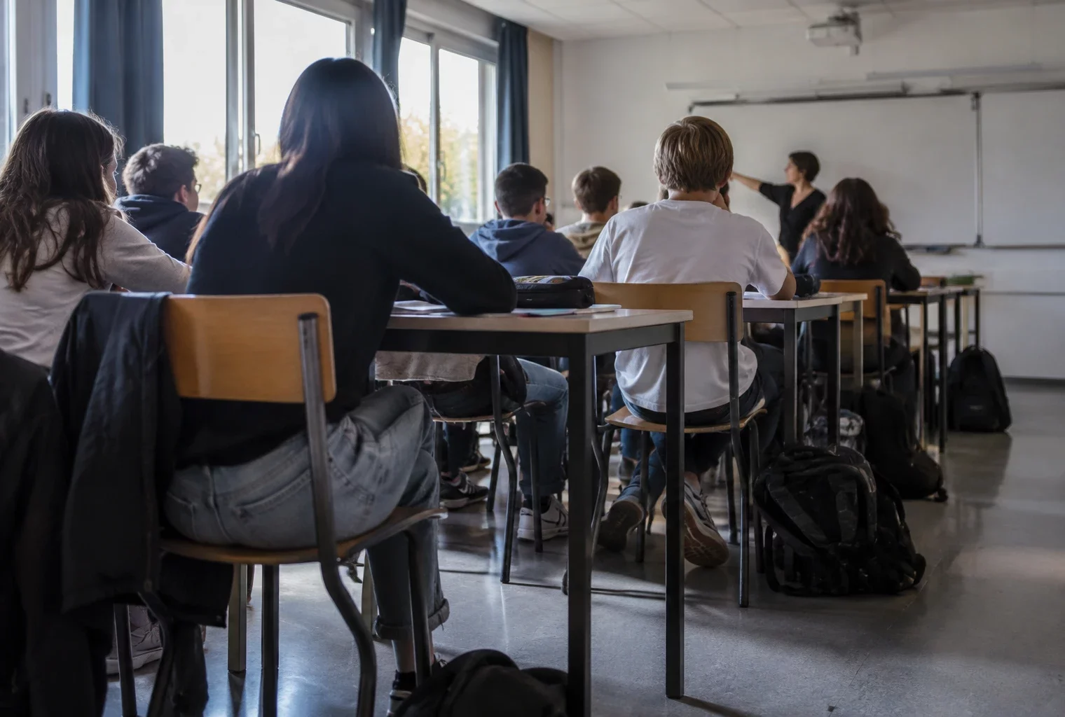 Tables rapprochées et chaises occupent une salle de classe de lycée, avec un tableau mural au fond.