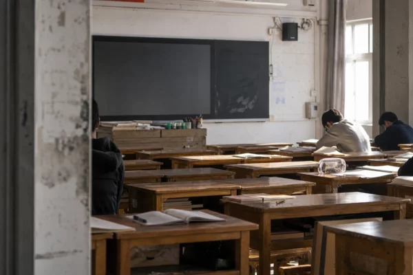 Classe de lycée en France, rangées de bureaux usés, tableau au fond et cahiers ouverts, lumière dure en contre-jour.