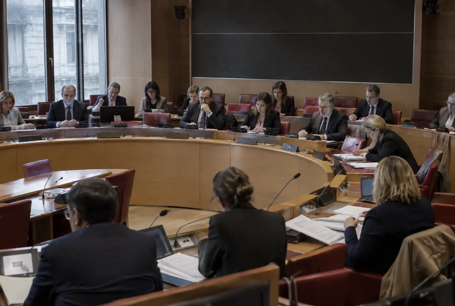 Pupitres équipés de microphones et dossiers ouverts dans une salle de commission parlementaire aux rangées de sièges vides.