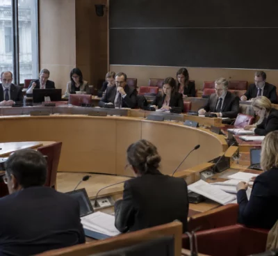 Pupitres équipés de microphones et dossiers ouverts dans une salle de commission parlementaire aux rangées de sièges vides.