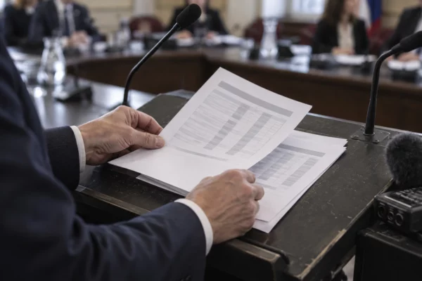 Des mains disposent des pages aux tableaux financiers abstraits sur un pupitre, dans une salle de réunion avec sièges et micros.