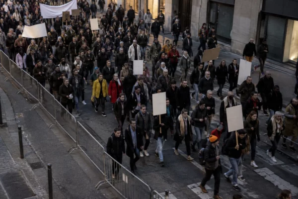 Un cortège de manifestants avance dans une rue bordée de façades, derrière des barrières métalliques et des pancartes illisibles.
