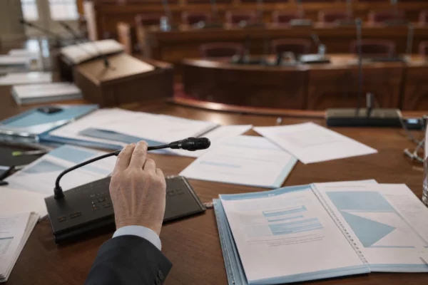 Réunion à la Cour des comptes : mains près d’un micro, dossiers budgétaires empilés et salle de conférence en arrière-plan.