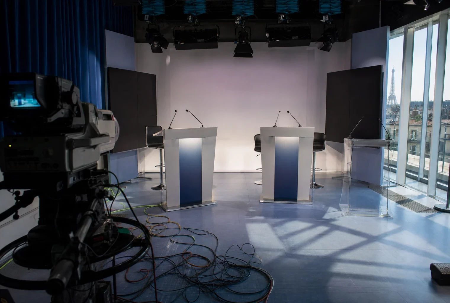 Plateau de débat télévisé à Paris, pupitres et micros au centre, câbles visibles, fond neutre sans logo.