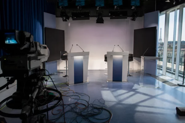 Plateau de débat télévisé à Paris, pupitres et micros au centre, câbles visibles, fond neutre sans logo.