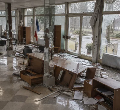 Salle municipale aux fenêtres endommagées, avec mobilier renversé et éclats de verre dispersés sur le sol carrelé.