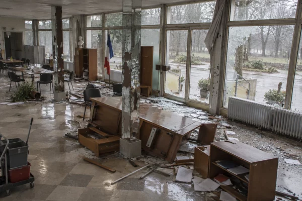Salle municipale aux fenêtres endommagées, avec mobilier renversé et éclats de verre dispersés sur le sol carrelé.