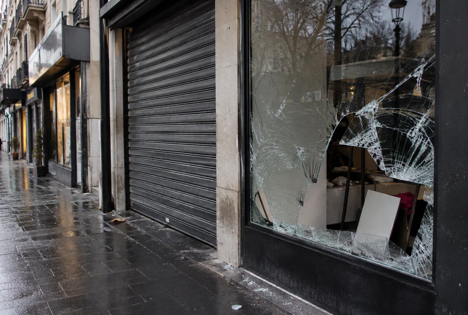 Devanture de commerce aux vitres fissurées et rideau métallique baissé, dans une rue bordée de boutiques.