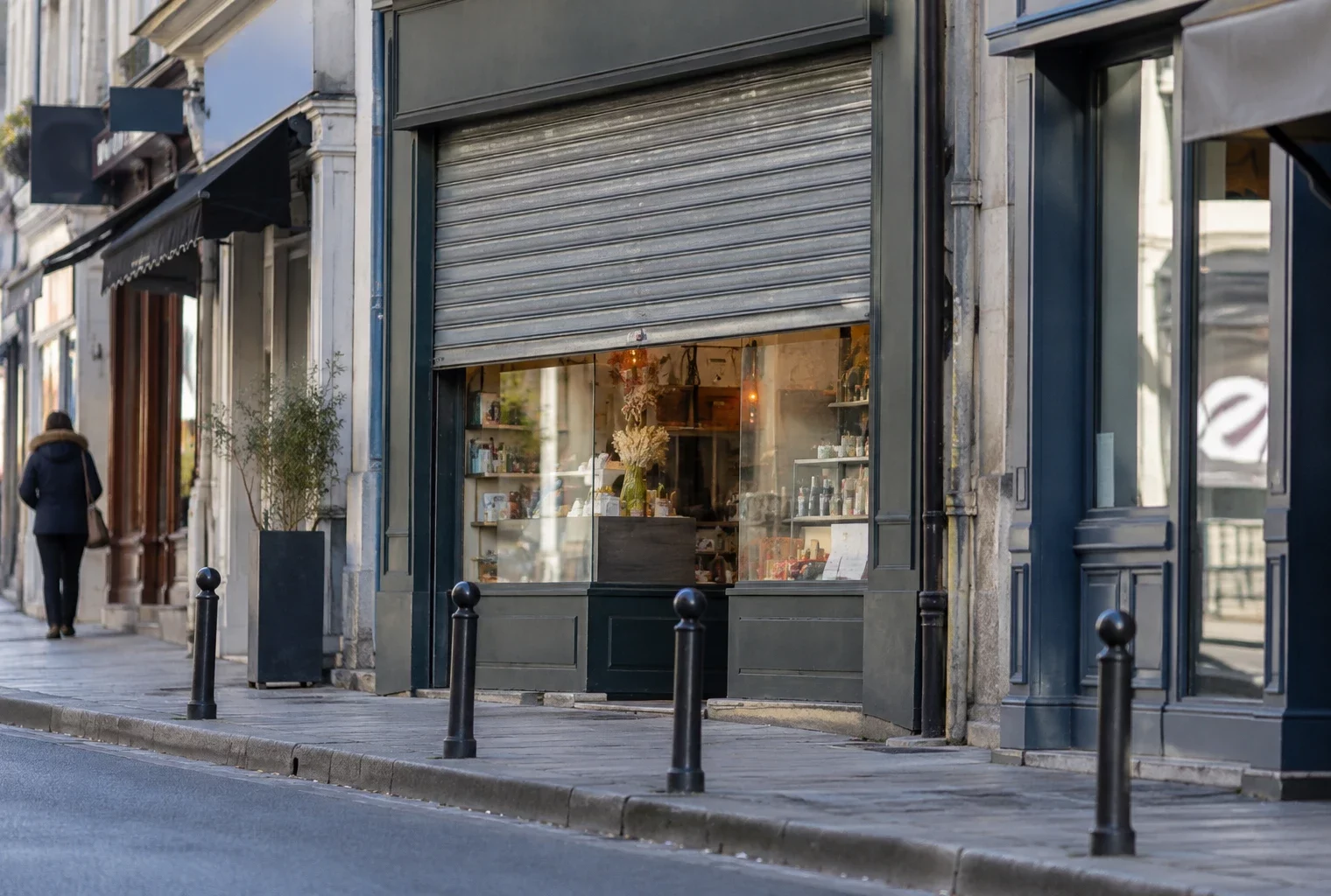 Devanture d’un commerce de quartier avec rideau métallique partiellement relevé, sur une rue bordée de boutiques.