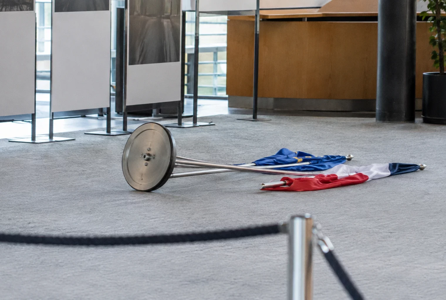 Support commun renversé et deux drapeaux tombés au sol dans un espace d’exposition aux panneaux sobres.