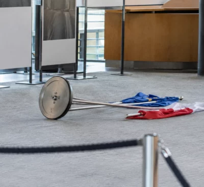 Support commun renversé et deux drapeaux tombés au sol dans un espace d’exposition aux panneaux sobres.