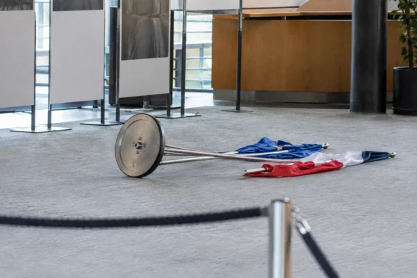 Support commun renversé et deux drapeaux tombés au sol dans un espace d’exposition aux panneaux sobres.