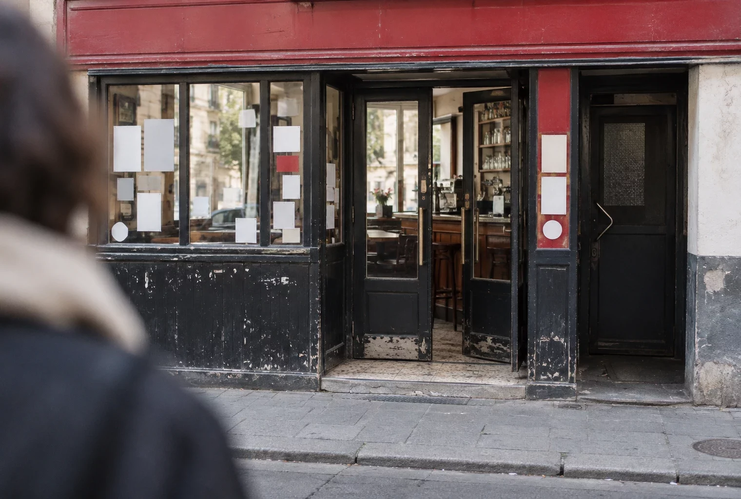 Entrée d’un bar parisien, porte vitrée ouverte sur un intérieur sobre avec comptoir et chaises, trottoir au premier plan.