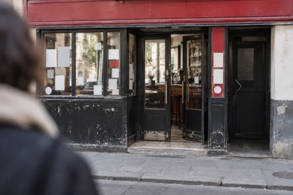 Entrée d’un bar parisien, porte vitrée ouverte sur un intérieur sobre avec comptoir et chaises, trottoir au premier plan.