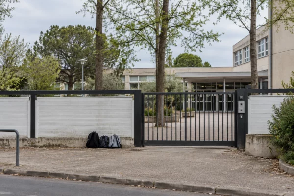 Un portail et une clôture devant un lycée, avec quelques sacs près du passage et des arbres au-dessus des bâtiments.