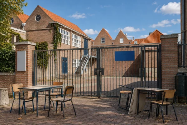 Quelques chaises et tables bloquent le portail d’un lycée, devant une cour pavée et une façade de briques aux fenêtres de classe.