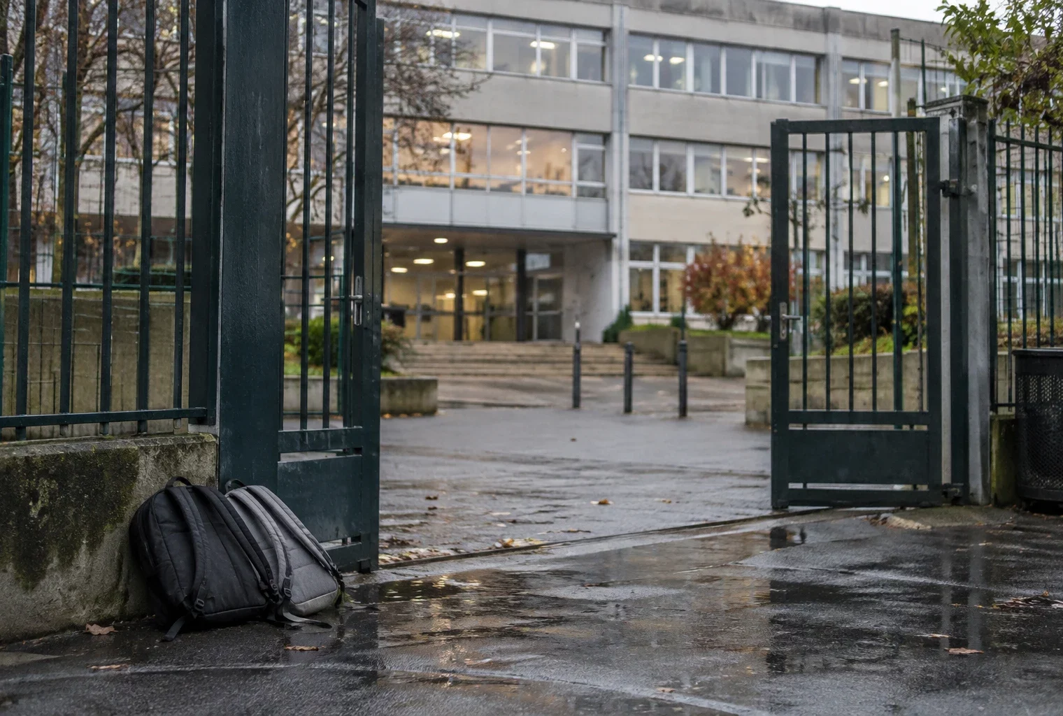 Grille d’entrée d’un lycée devant une façade scolaire, avec quelques sacs posés près du passage et des fenêtres de classe.