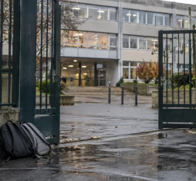 Grille d’entrée d’un lycée devant une façade scolaire, avec quelques sacs posés près du passage et des fenêtres de classe.