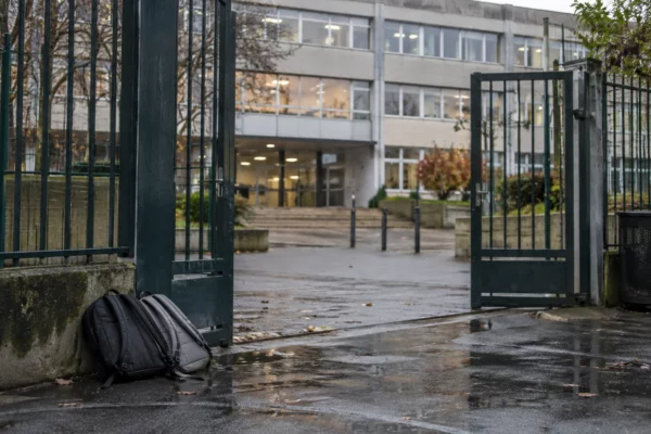 Grille d’entrée d’un lycée devant une façade scolaire, avec quelques sacs posés près du passage et des fenêtres de classe.