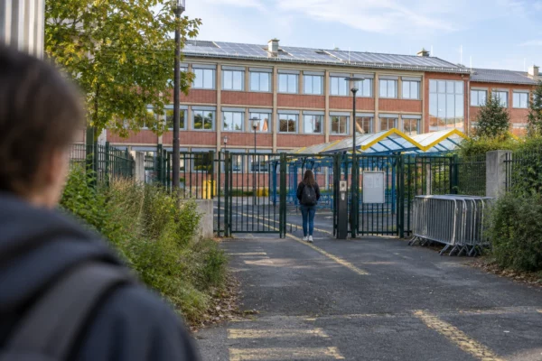 Entrée d’un lycée en briques avec portail métallique, barrières rangées sur le côté et passage couvert devant les fenêtres.