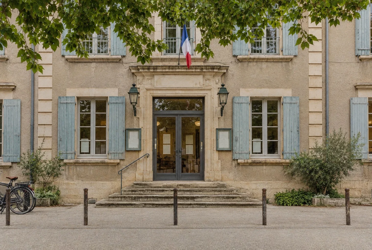 Entrée vitrée d’un bâtiment municipal en pierre, avec seuil usé, vélos et bornes devant la façade sous un ciel couvert.