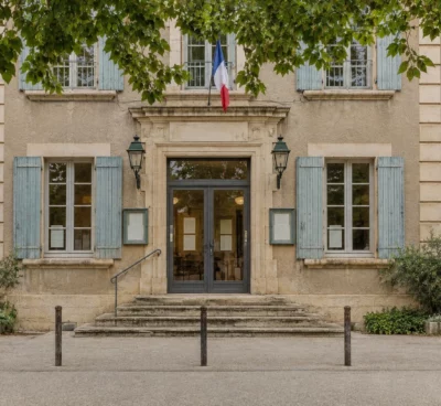 Entrée vitrée d’un bâtiment municipal en pierre, avec seuil usé, vélos et bornes devant la façade sous un ciel couvert.