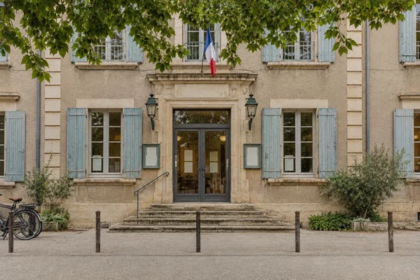 Entrée vitrée d’un bâtiment municipal en pierre, avec seuil usé, vélos et bornes devant la façade sous un ciel couvert.
