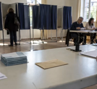 Une enveloppe et des bulletins sur une table de vote, avec des isoloirs et des registres visibles dans la salle.