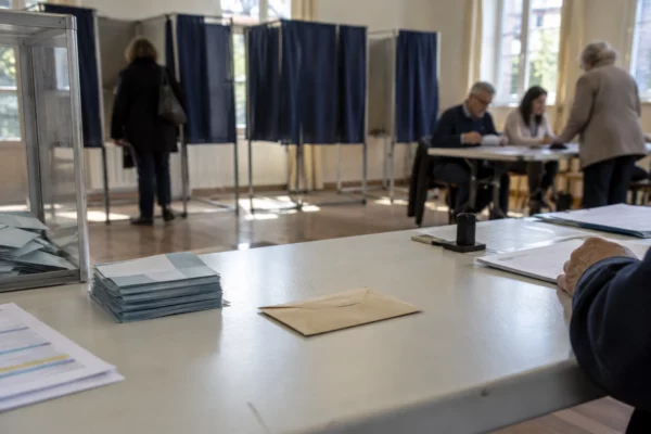 Une enveloppe et des bulletins sur une table de vote, avec des isoloirs et des registres visibles dans la salle.
