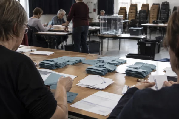 Des enveloppes électorales ouvertes et regroupées sur une table, avec des feuilles de comptage et des bacs de tri en arrière-plan.