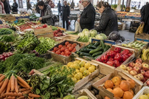 Des fruits et légumes frais sont disposés dans des cagettes sur un étal de marché, avec d’autres stands en arrière-plan.