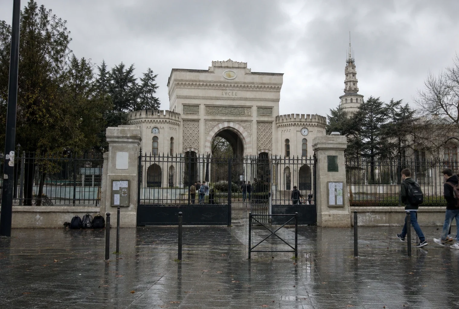 Portail métallique d’un lycée français sous la pluie, trottoir mouillé, façade scolaire en arrière-plan et ambiance grise.