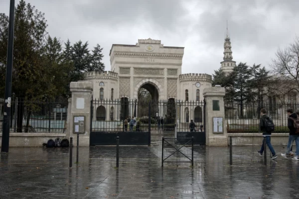 Portail métallique d’un lycée français sous la pluie, trottoir mouillé, façade scolaire en arrière-plan et ambiance grise.