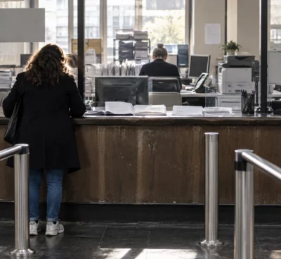 Un dossier administratif ouvert et des formulaires vierges sur le comptoir usé d’un guichet d’accueil, avec des postes de traitement en arrière-plan.