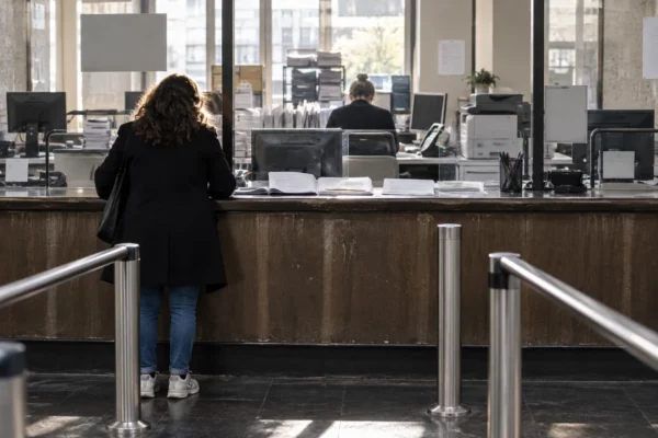 Un dossier administratif ouvert et des formulaires vierges sur le comptoir usé d’un guichet d’accueil, avec des postes de traitement en arrière-plan.