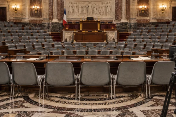Dans un hémicycle parlementaire vide, des pupitres équipés de microphones font face aux rangées de sièges et à la tribune.