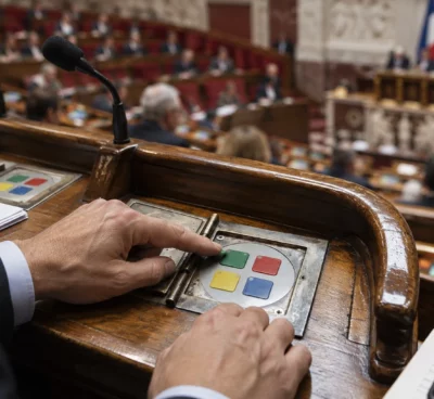 Des mains actionnent un boîtier de vote sur un pupitre usé, avec les rangées de sièges de l’hémicycle en arrière-plan.