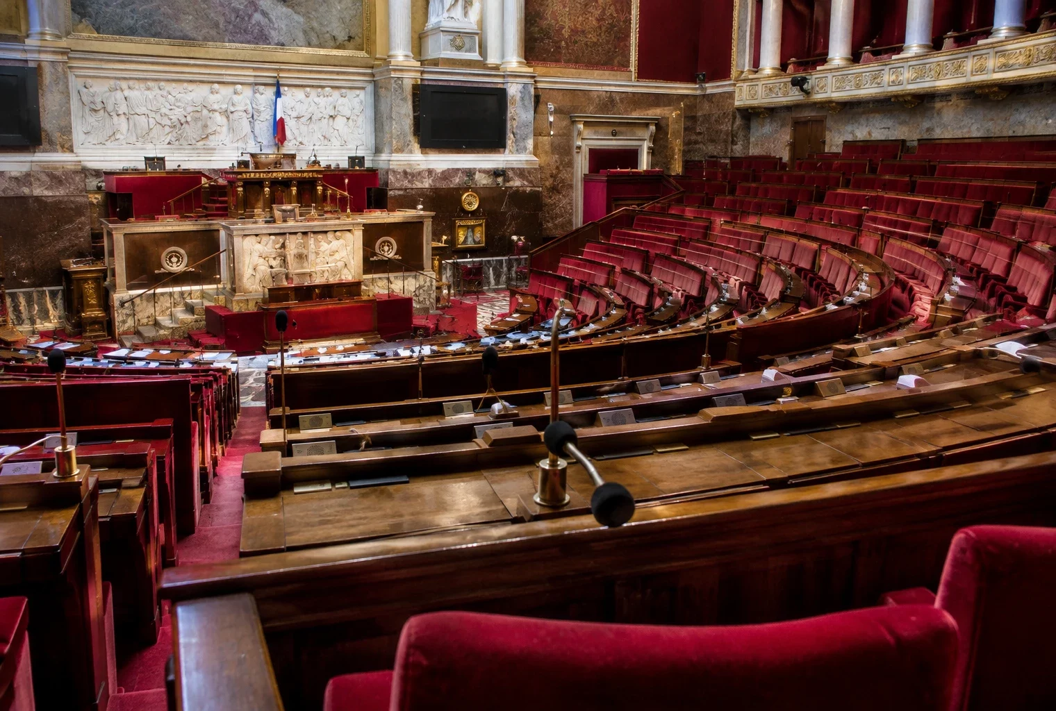 Pupitres équipés de microphones dans un hémicycle parlementaire, avec des rangées de sièges et une tribune au fond.