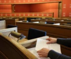 Gros plan sur des mains anonymes dans l’hémicycle vide du Landtag, entre pupitres en bois, micros et lumière matinale.