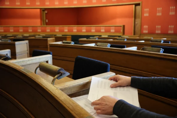 Gros plan sur des mains anonymes dans l’hémicycle vide du Landtag, entre pupitres en bois, micros et lumière matinale.