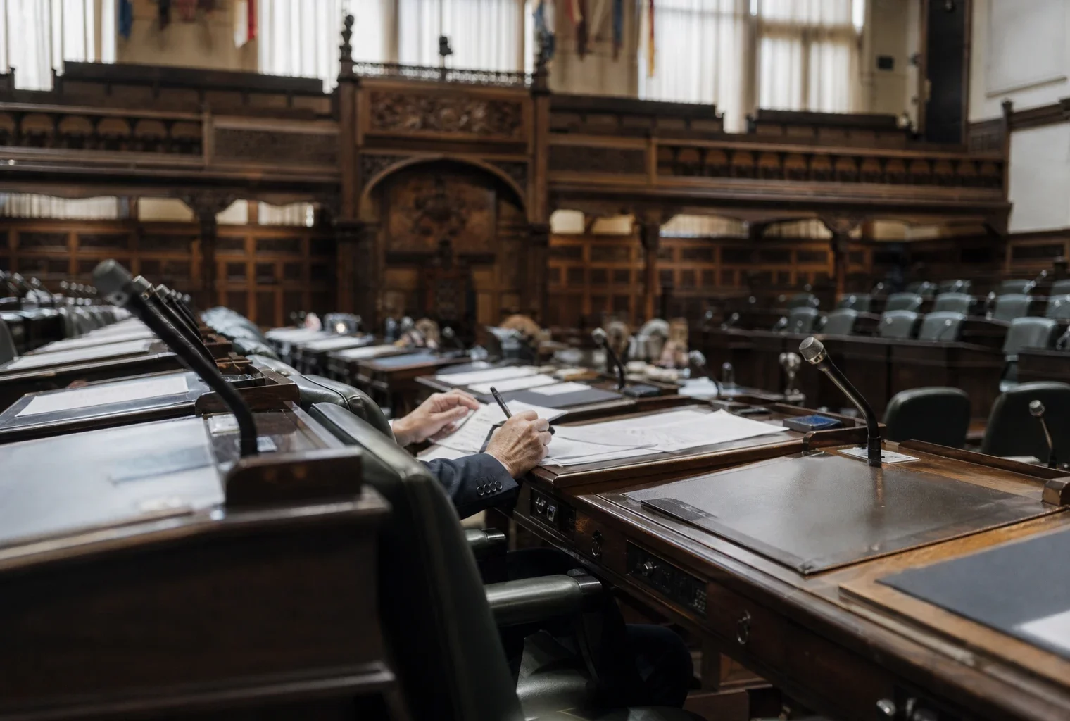 L’hémicycle du Sénat vu en biais, avec pupitres, micros et mains anonymes sur un dossier au premier plan.