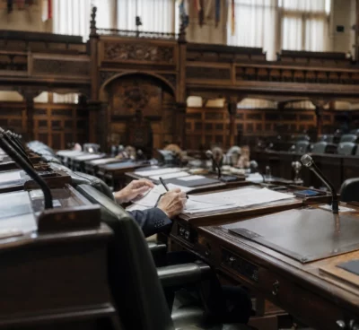 L’hémicycle du Sénat vu en biais, avec pupitres, micros et mains anonymes sur un dossier au premier plan.