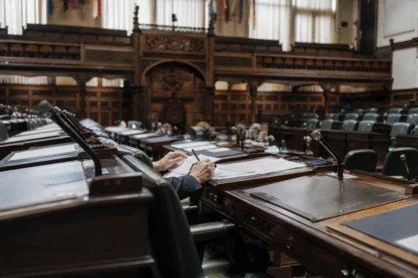 L’hémicycle du Sénat vu en biais, avec pupitres, micros et mains anonymes sur un dossier au premier plan.