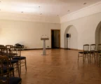 Salle de cérémonie aux Invalides, rangées de chaises, pupitre et murs clairs sous une lumière rasante.