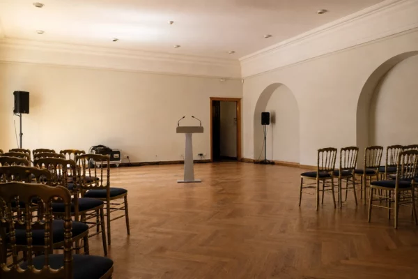Salle de cérémonie aux Invalides, rangées de chaises, pupitre et murs clairs sous une lumière rasante.