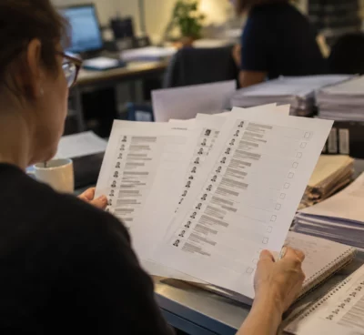 Des feuilles de questionnaire et des listes de candidats imprimées sont posées sur un bureau, avec d’autres documents empilés en arrière-plan.