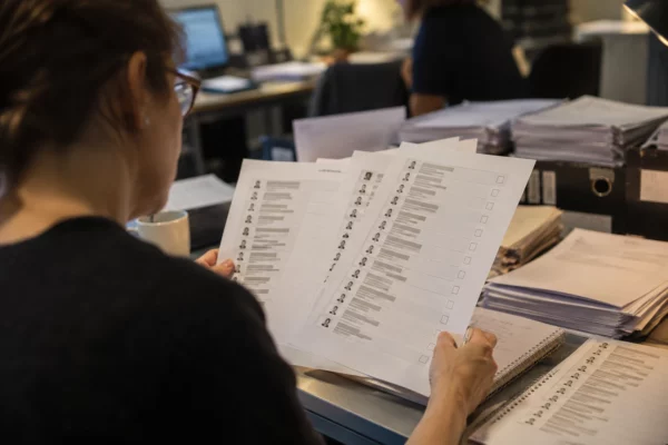 Des feuilles de questionnaire et des listes de candidats imprimées sont posées sur un bureau, avec d’autres documents empilés en arrière-plan.
