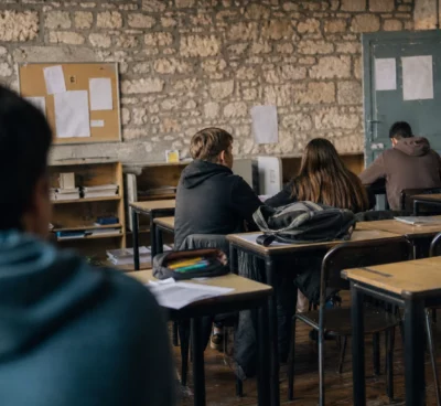 Intérieur de lycée public usé, rangées de tables, murs marqués et portes de salle floues dans une lumière douce.