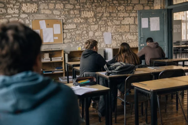 Intérieur de lycée public usé, rangées de tables, murs marqués et portes de salle floues dans une lumière douce.