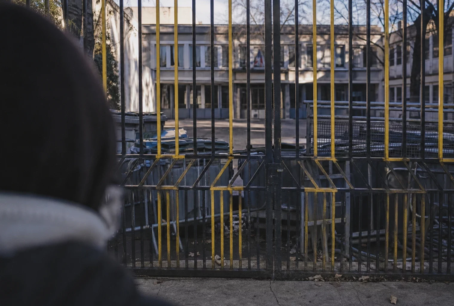 Portail de lycée fermé par des poubelles et barrières, vu de trois quarts, sous un soleil d’hiver bas.
